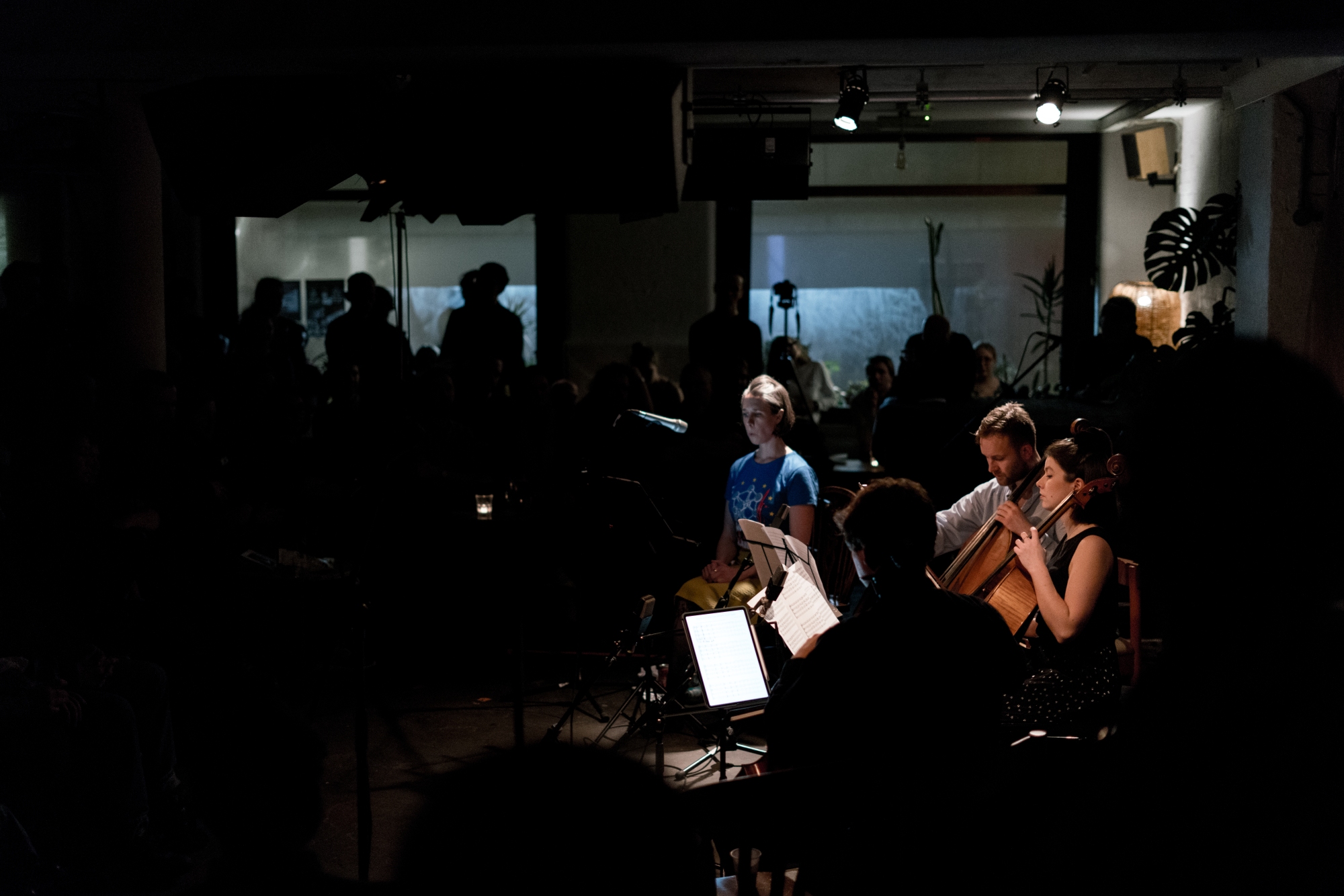 Photograph of a singer and three cellists performing in a dark and crowded room.