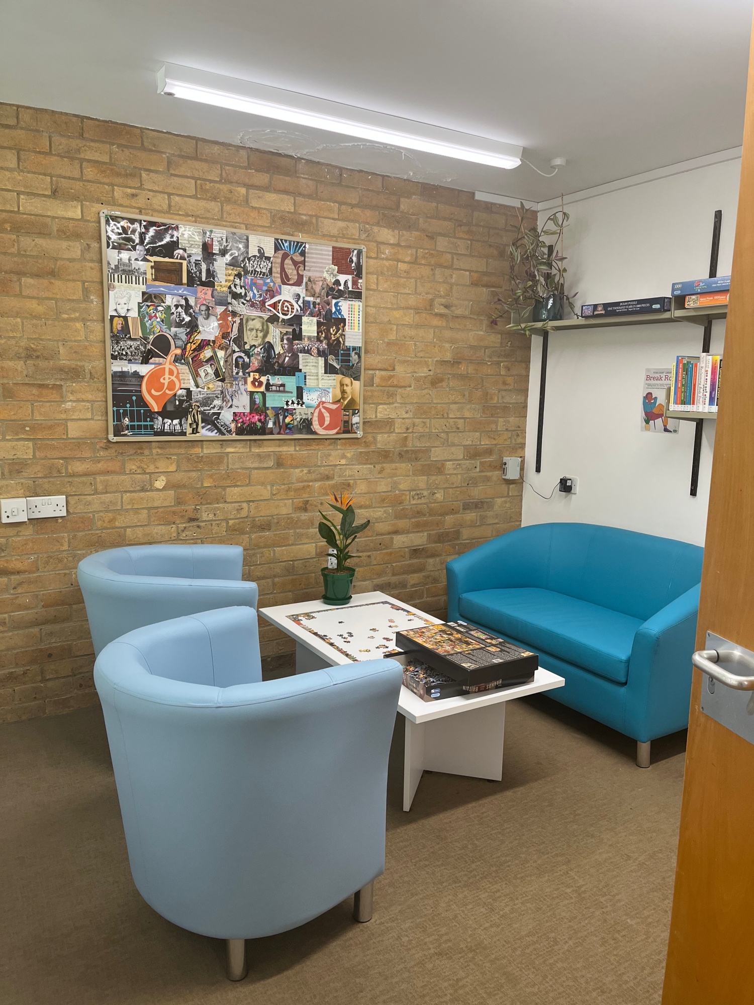 Photograph of library break room. Two light blue armchairs and a dark blue sofa surround a white coffee table with an unfinished puzzle and a potted plant on top. There is a large pinboard decorated with photos of composers on the brick wall at the back, and shelving with books on the right wall. 