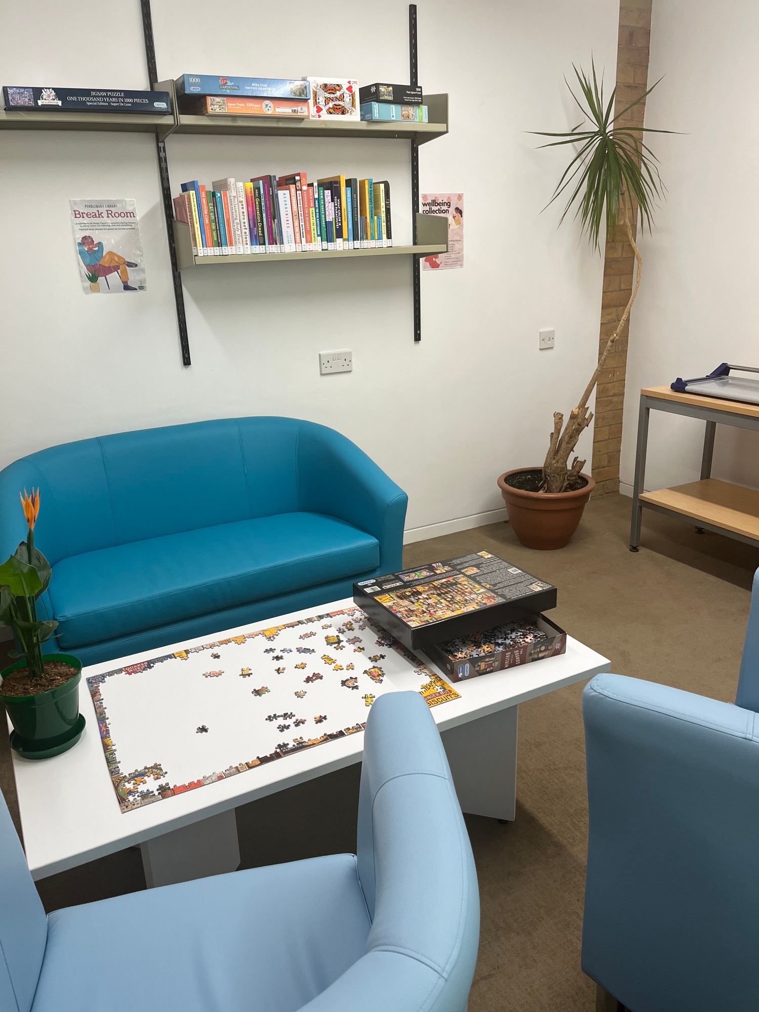 Photograph of library break room, featuring two pale blue arm chairs opposite a medium blue sofa, There is a white coffee table in between, with an unfinished puzzle and a potted plant on top. On the wall behind there is a shelf with books and board games. To the left is a large potted plant.
